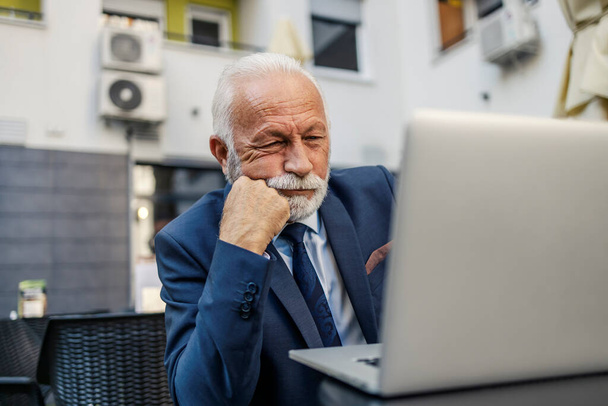 A tired senior businessman is sitting at the terrace of the cafe with a hand under his chin and looking at his laptop. He is reading the report on the laptop. A businessman working on a laptop at a cafe. - Photo, Image