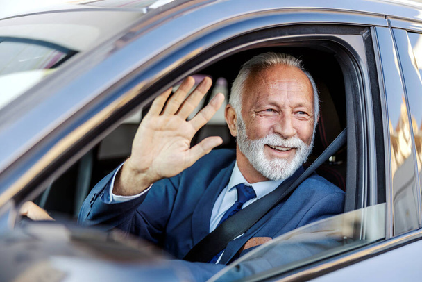 Ένας χαμογελαστός ανώτερος επιχειρηματίας με κοστούμι οδηγεί το αυτοκίνητό του και χαιρετάει. Φιλικός επιχειρηματίας χαιρετά άλλους οδηγούς. - Φωτογραφία, εικόνα