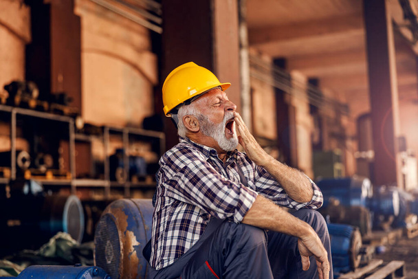 Ein müder Senior sitzt neben den Maschinen und gähnt. Nach harter Arbeit muss er schlafen. Ein schläfriger Arbeiter in der Fabrik. - Foto, Bild