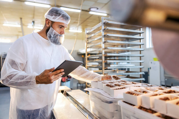 A food inspector in a sterile white uniform is holding the tablet and looking at collected cookies. Food check is important if we want quality and healthy food. - Photo, Image