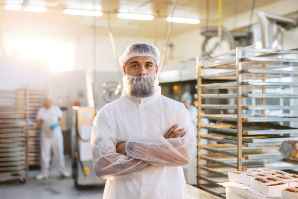 Um supervisor de fábrica de alimentos bem sucedido em um uniforme estéril branco está de pé na fábrica com os braços cruzados. Ele está muito satisfeito com a qualidade e quantidade de produtos. - Foto, Imagem