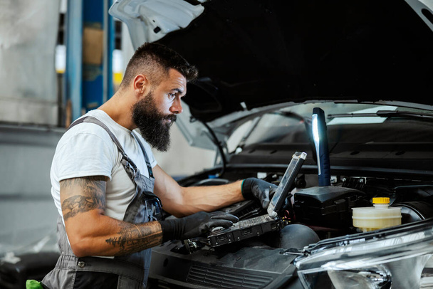Vedle rozbitého auta stojí vousatý automechanik v pracovní uniformě a k údržbě auta používá speciální notebook. Auto-mechanik v dílně opravuje auto - Fotografie, Obrázek