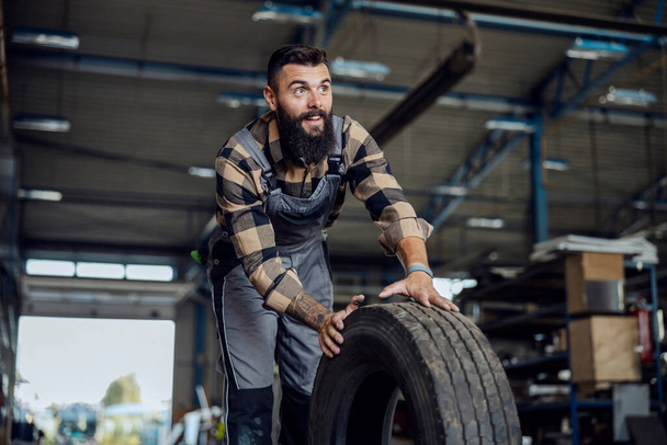 A motivated auto-mechanic in working uniform rolling tire and preparing to change it. Auto-mechanic in a workshop with tire - Photo, image