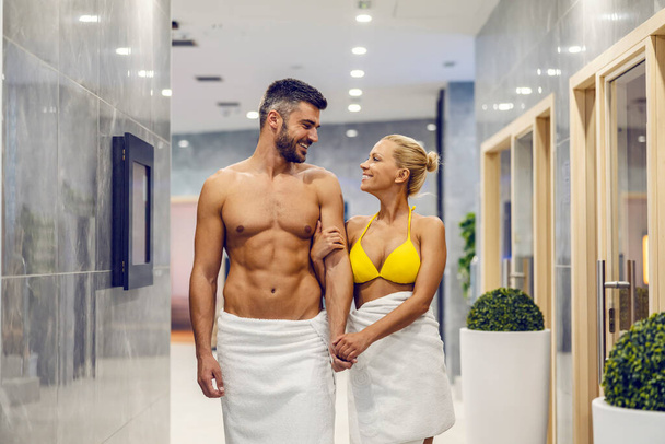 Una encantadora pareja atractiva en toallas está cogida de la mano y caminando en el centro de spa. El tratamiento del cuerpo sano es el es importante como es amar a sí mismo ya su pareja. Una pareja feliz en el spa. - Foto, imagen