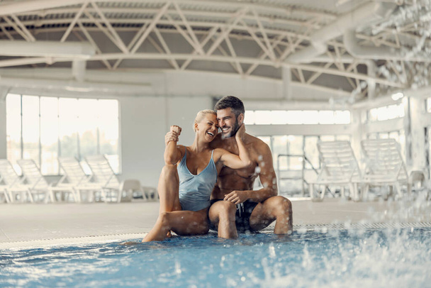 Šťastný pár sedí u bazénu s bublající vodou, směje se a objímají v lázních a wellness centru. Pár sedící vedle termální vody - Fotografie, Obrázek