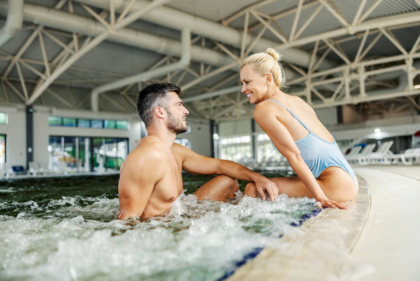 温泉センターでお湯でプールの隣に座って男とイチャイチャする幸せなセクシー女性。スパセンターのカップル. - 写真・画像
