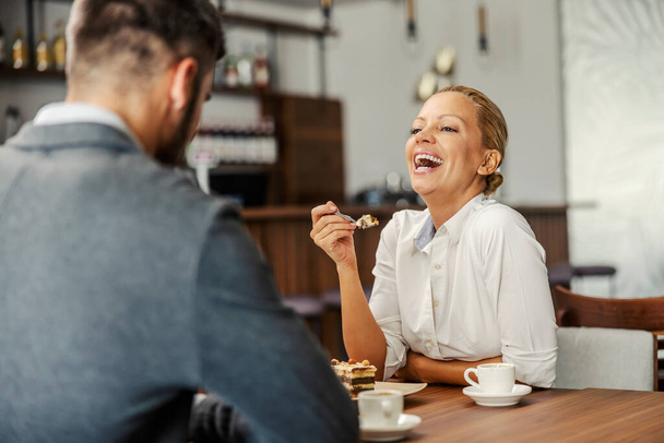 Mutlu, zarif bir kadın bir restoranda kocasıyla oturuyor, esprilerine gülüyor ve lezzetli pastalar yiyor. Restoranda tatlı yiyen bir çift. - Fotoğraf, Görsel