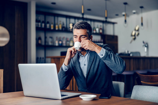 A businessman is sipping his coffee while looking at the laptop. He finishes his work on a laptop in working friendly cafe. A businessman drinks coffee and finishing his work at the cafe. - Photo, Image