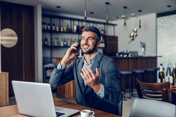A friendly busy man is sitting in the working friendly cafe and having a phone call with colleagues. There is a laptop on a table. A businessman talking on the phone in a cafe - Foto, Bild