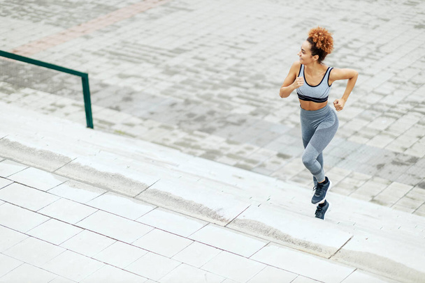 Das Mädchen rennt. Ein Mädchen mit lockig gebundenen Haaren in grauer Sportbekleidung mit einem Lächeln im Gesicht läuft von der Treppe herunter und schaut von der Seite. Fitness und gute körperliche Verfassung - Foto, Bild