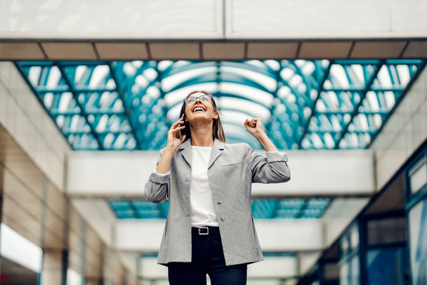 Mädchen und gute Nachrichten. Eine Frau in lässiger Jacke und Brille hält sich das Telefon ans Ohr und hüpft vor Freude über die gute Nachricht, die ihr am Telefon im Geschäftssaal überbracht wurde. Erfolg bei der Arbeit - Foto, Bild