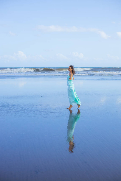Young woman walking barefoot on empty beach. Full body portrait. Slim Caucasian woman wearing long dress. Water reflection. Summer sun light. Blue sky. Vacation in Asia. Travel concept. Bali Indonesia - Foto, Imagem