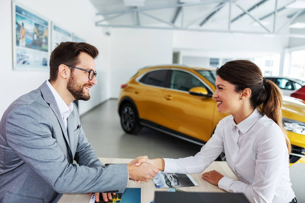 Gülümseyen, dost canlısı bir araba satıcısı bir müşteriyle masada oturuyor ve el sıkışıyor. Kadın yeni bir araba aldı ve çok memnun.. - Fotoğraf, Görsel