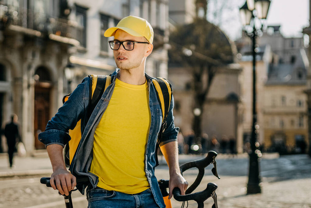 Bisiklette oturan mutlu bir teslimatçı. O, sarı elbise giyiyor. O, omuzlarında sarı bir sırt çantası taşıyor. Eski şehrin merkezi.. - Fotoğraf, Görsel