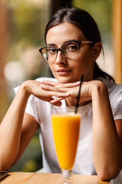 Barda poz veren ve kameraya bakan genç bir kadının portresi. Önünde bir bardak taze portakal suyu var. Barda bir kız - Fotoğraf, Görsel