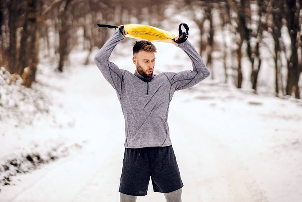 Fit αθλητής κάνει ασκήσεις με βάρη, ενώ στέκεται σε χιονισμένο μονοπάτι στη φύση το χειμώνα. Χειμερινή φυσική κατάσταση, bodybuilding, υγιεινές συνήθειες - Φωτογραφία, εικόνα