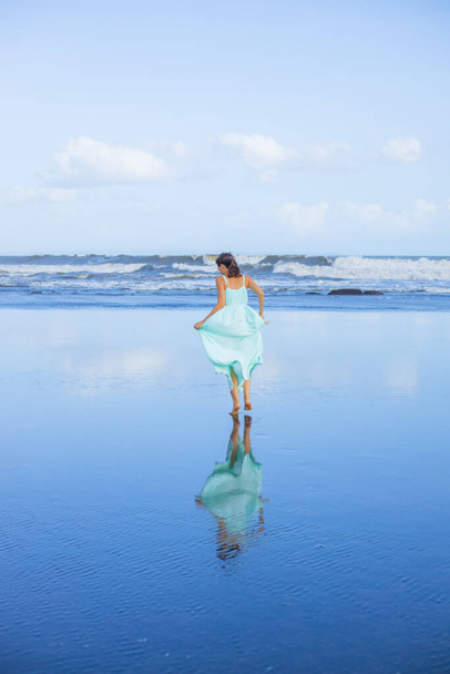 Nuori nainen kävelee paljain jaloin tyhjällä rannalla. Koko vartalon muotokuva. Hoikka valkoihoinen nainen yllään pitkä mekko. Näkymä takaa. Vedenheijastus. Sininen taivas. Loma Aasiassa. Matkakonsepti Bali, Indonesia - Valokuva, kuva