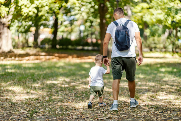 Familie wandeling in het bos. Een volwassen man loopt en houdt de hand vast van een kleine jongen die dezelfde kleren heeft als zijn vader. Vader en zoon, gedeelde momenten van een prachtige zomerse dag in de natuur - Foto, afbeelding