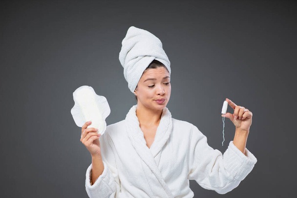 Azon gondolkodom, hogy törlőkendőt vagy tampont használok. Egy határozatlan nő fürdőköpenyben és egy törölközővel a fején higiéniai tárgyakat tart a menstruációja alatt. Női termék, kozmetikai termékek - Fotó, kép