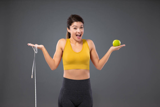 Nutrition and healthy habits. A smiling woman in sportswear stands in front of a gray wall and holds an apple in one hand and a weight measuring tape in the other. Low-calorie food and training - Photo, Image