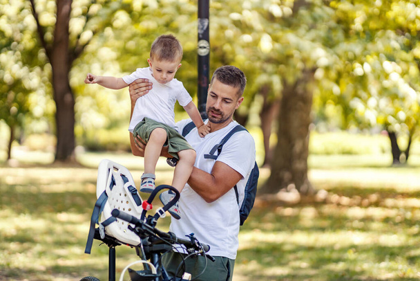 Turvallinen pyöräily ja huoleton perhe pyöräilemässä. Aikuinen uros laittaa lapsen pyöräkoriin ja valmistautuu turvalliseen ajoon. Pyöräily ja aktiivinen perheviikonloppu puistossa kesällä - Valokuva, kuva