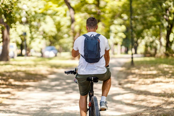 Man and bicycles. An adult male dressed in casual short-sleeved T-shirt and olive-green shorts with a backpack riding bicycles in the park Back shot, sports routine and hobby during summertime in park - Photo, Image