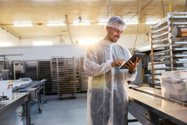 Een tevreden voedselfabriek inspecteur in een steriel uniform gebruikt een tablet om de kwaliteit en kwantiteit van de producten te controleren. Hij staat naast een rij. Hij is erg blij met de resultaten.. - Foto, afbeelding