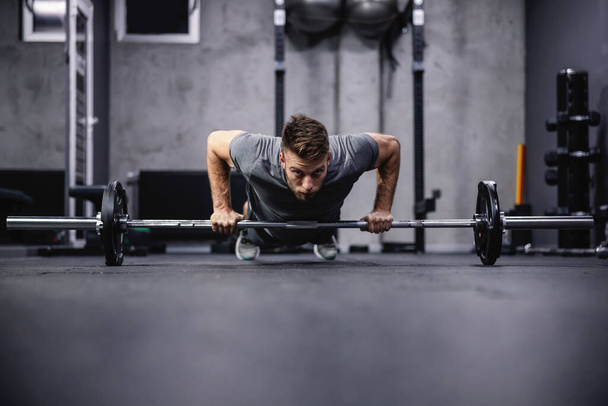 Push-ups and muscle strengthening. An attractive and muscular man does push-ups leaning his hands on a barbell. A man in sportswear pumps his arm muscles in a modern gym. Sport, fitness challenge - Photo, Image