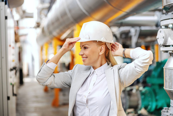 Getting ready for work. Smiling blond businesswoman in formal wear putting the protective helmet on head and preparing to walk around the power plant. A woman in the business world - Photo, Image