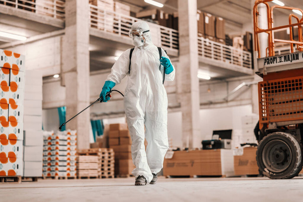 Koronavirüsle ilgili ciddi bir durum. COVID19 'un ve fabrika deposundaki temizlik yerinin koruyucu beyaz takım elbiseli biri tarafından kapatılması. Temizlik spreyi, koruyucu kalmak, korona durumu - Fotoğraf, Görsel