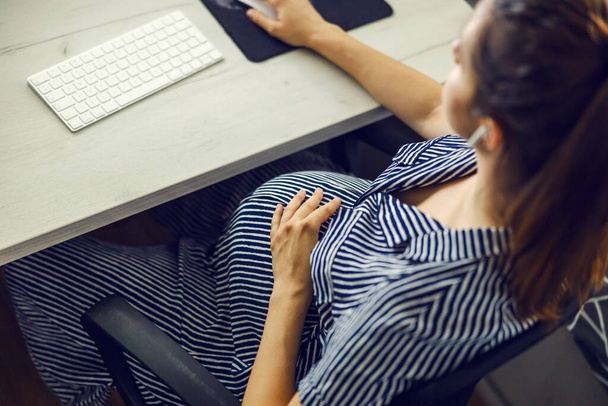 妊娠中の女性とコンピューター家庭局妊娠中のビジネスウーマン。巨大な胃を持つ女性は、彼女の耳にヘッドフォンでコンピュータの前に座っており、オンラインレッスンのためのマウスを使用しています - 写真・画像
