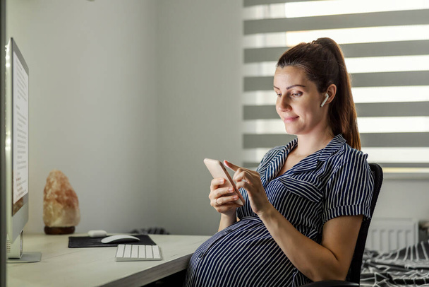 Femme enceinte utilisant un téléphone au bureau à la maison. Une femme enceinte avec succès en robe rayée s'assoit devant un ordinateur avec un casque dans les oreilles et utilise le téléphone. En ligne, écouter de la musique - Photo, image