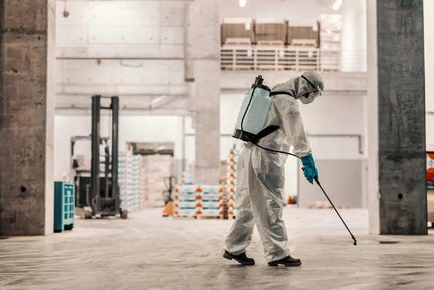 Person in a white protective suit refreshing and cleaning a fabric warehouse indoor space. Fight against COVID 19, coronavirus life, stay healthy, keep social distance, stop spreading corona virus - Photo, Image