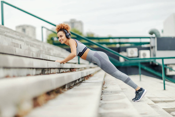 Young sportswoman with healthy habits is listening to the radio while doing push-ups on the stairs in an urban district. - Photo, Image