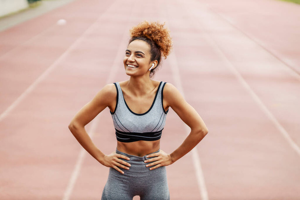 Smile and training. A girl with curly tied hair dressed in gray sportswear with headphones in her ears holds her hands on her hips while listening to relaxing music and laughs for a successful workout - Photo, Image