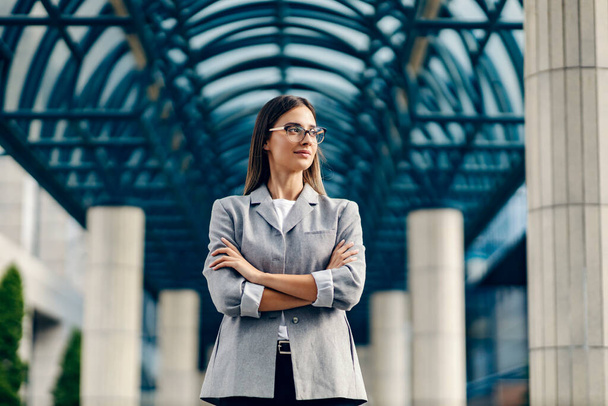 Een jonge, succesvolle vrouwelijke baas gekleed slim casual staat in het business center met de armen gekruist en op zoek naar een nieuwe kans. - Foto, afbeelding