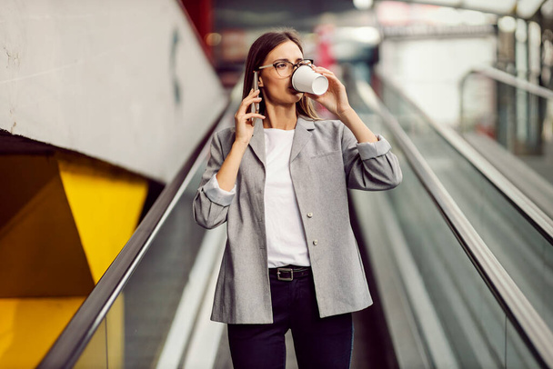 Egy fiatal, elfoglalt üzletasszony megy le a mozgólépcsőn és issza a kávéját, miközben egy fontos telefonbeszélgetést folytat egy ügyféllel.. - Fotó, kép