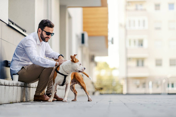 A businessman dressed smart casual sitting on the sidewalk with and petting his dog. On his lunch break, he chose to walk his dog. - Foto, Imagem