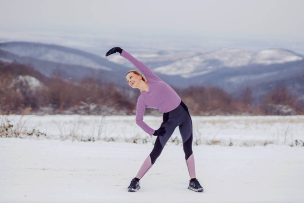 Sıska bir sporcu kadın, karlı kış gününde doğada ısınma ve esneme egzersizleri yapıyor. Sağlıklı alışkanlıklar, kış sporu - Fotoğraf, Görsel