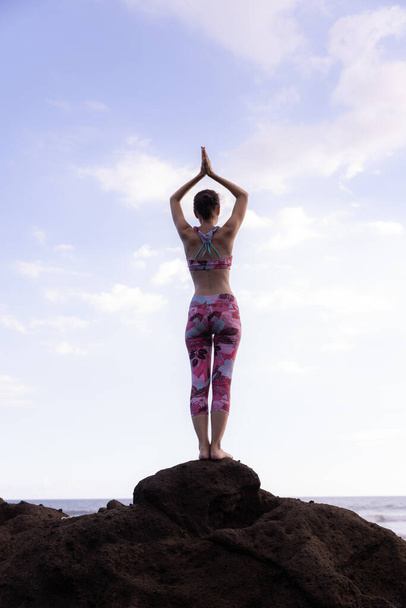 Woman standing on the rock, practicing yoga. Young woman raising arms with namaste mudra at the beach. Blue sky background. Yoga retreat in Bali. View from back. Vertical layout. Copy space. Indonesia - Photo, Image