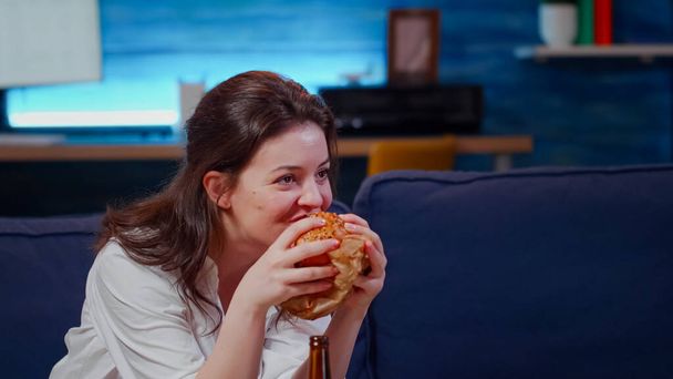Young person laughing at TV while eating hamburger - Foto, Imagen