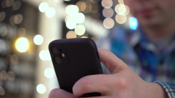 A young man is sitting at a table in a cafe in the evening and texting on the phone. A man with a phone in his hands on the background of garlands. Close-up. - Footage, Video