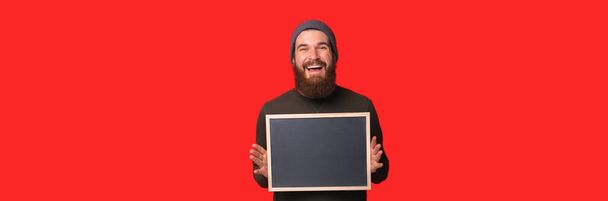 Banner tamaño foto de sonriente joven barbudo sosteniendo pizarra vacía sobre fondo rojo - Foto, Imagen