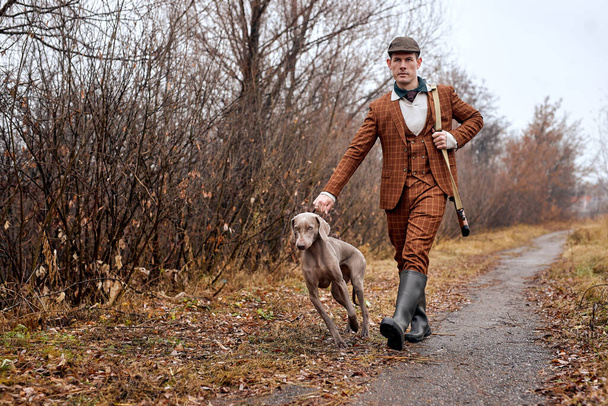male hunter in suit ready to hunt, holding gun and walking with dog in forest. hunting - Fotoğraf, Görsel