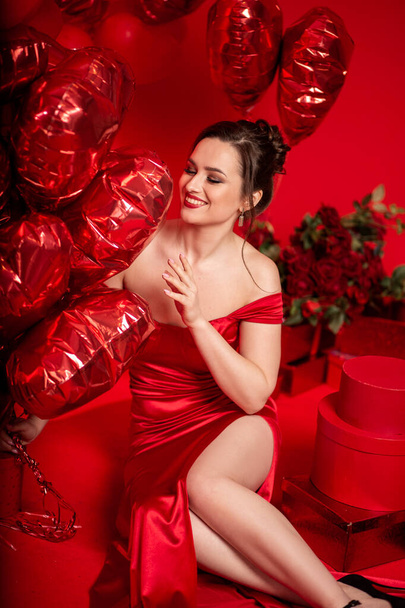 Beautiful young woman in red evening dress posing over red background with big heart shape balloons. - Valokuva, kuva