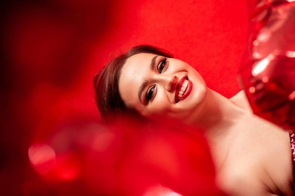 Beautiful young woman in red evening dress posing over red background with big heart shape balloons. - Foto, imagen
