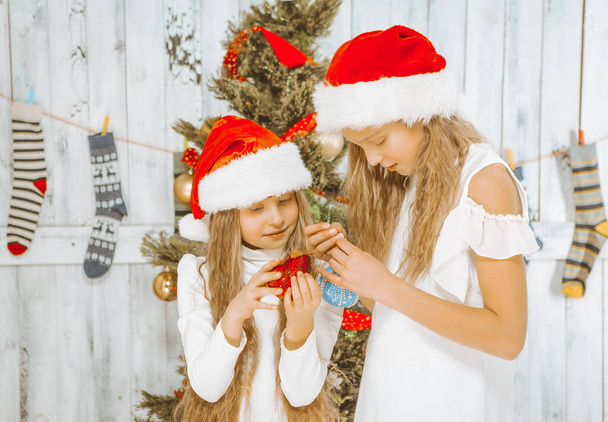 Las adolescentes con vestidos blancos y sombreros rojos admiran las decoraciones navideñas en sus manos. Preparándose para Navidad, Nochebuena. Primer plano. Fondo de madera - Foto, imagen