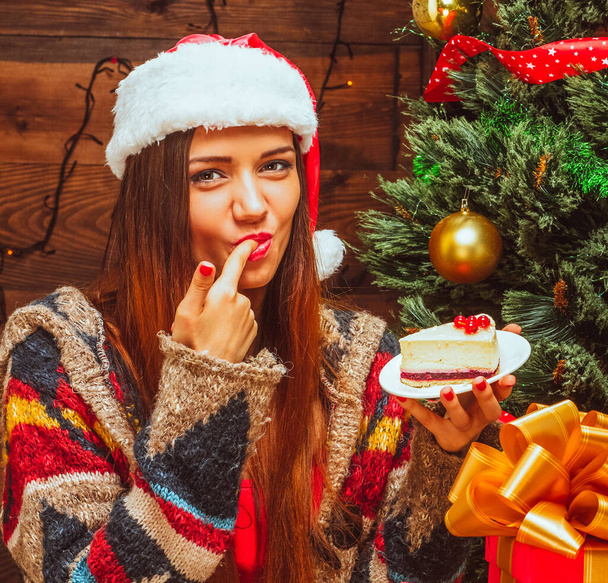 bright lady holding a plate with a slice of cake and tasting it, finger in the mouth, Christmas Eve, Christmas dinner, Christmas cake, Christmas Tree, Christmas Gifts, Wood Background. Close-up - Foto, afbeelding