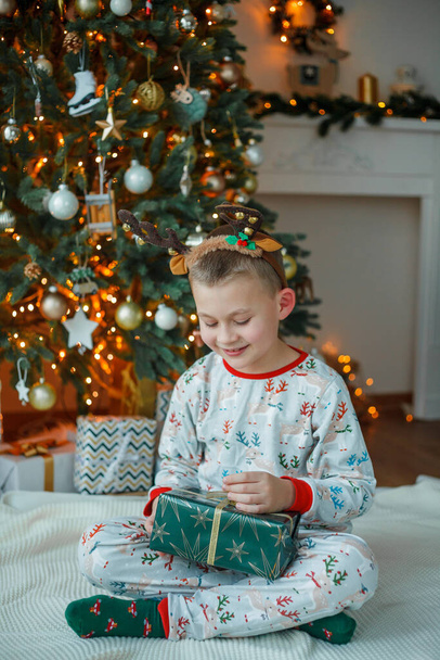 Adorable 7 year old boy in pajamas opens gift box near the Christmas tree. New Year's lights. Winter holidays. Cozy home. - Foto, Imagen