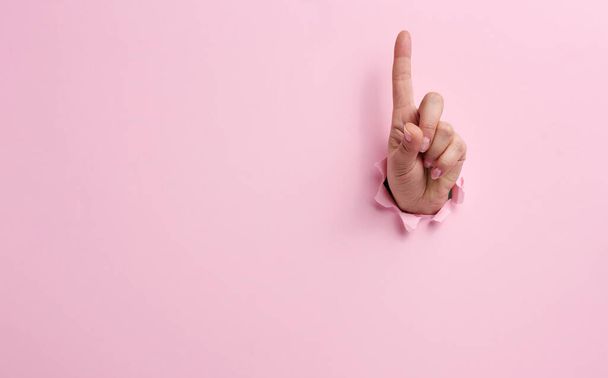 mano femenina que sobresale de un agujero roto en un fondo de papel rosa, gesto de atención. Lugar para una inscripción - Foto, imagen